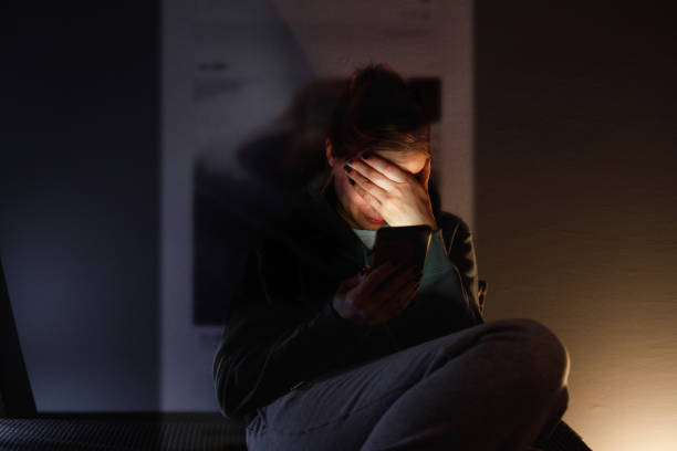 A 34 year old woman is sitting in the dark room and using her smartphone.
